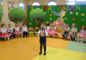 Chłopiec trzyma w ręku mikrofon, recytuje wiersz. Wokół chłopca siedzą dzieci.